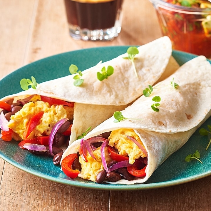 A plate of two rolled tortillas filled with scrambled eggs, black beans, red peppers, and onions, garnished with small green sprouts, accompanied by a glass of dark coffee and a small bowl of salsa, all set on a wooden table.